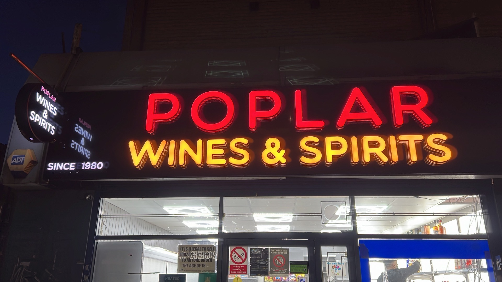 The red and gold neon sign of Poplar Wines & Spirits glowing at night above the shopfront on Poplar High Street, E14