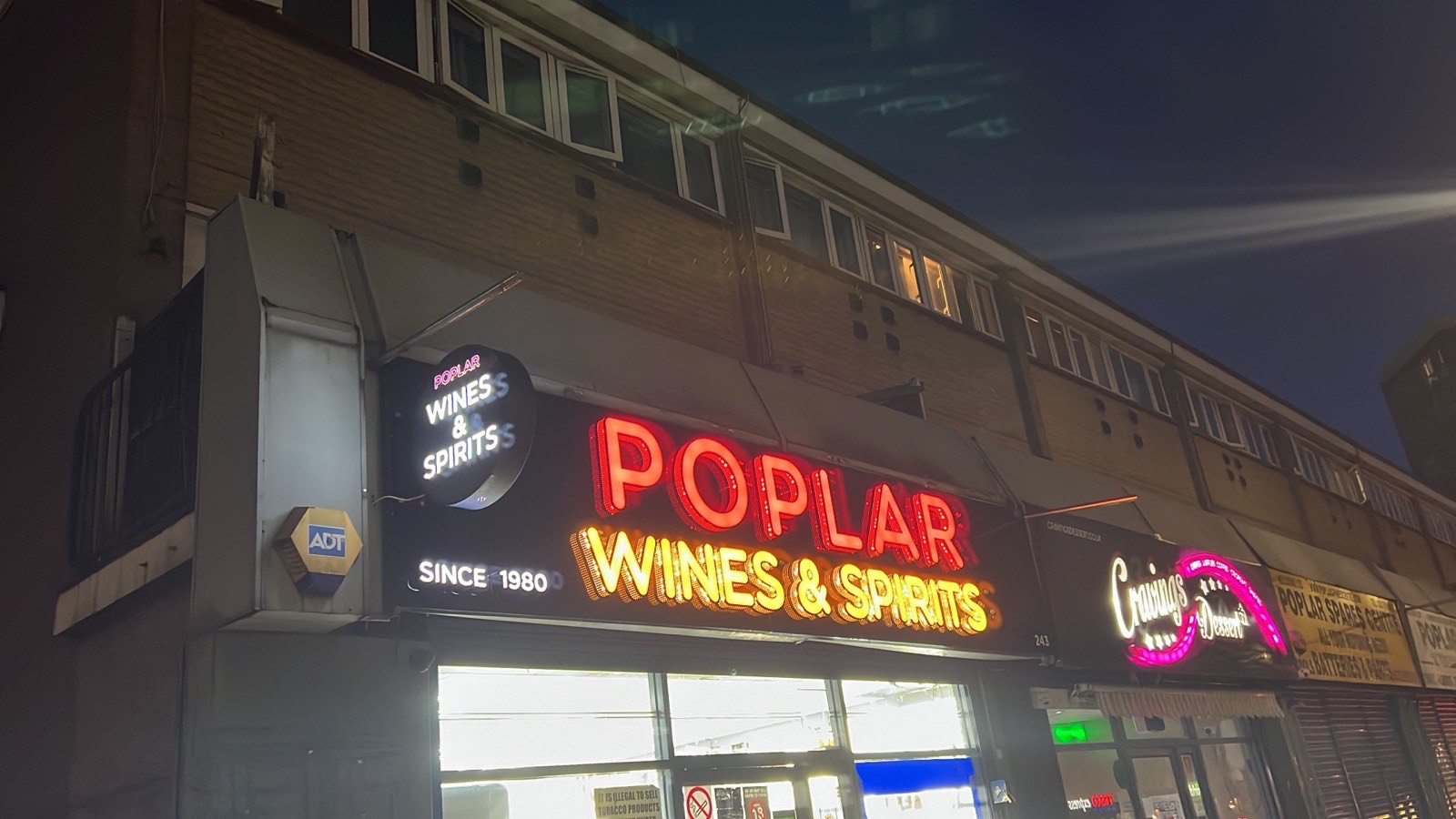 The Poplar Wines & Spirits shopfront on the corner of Poplar High Street at night, its neon sign reading Since 1980